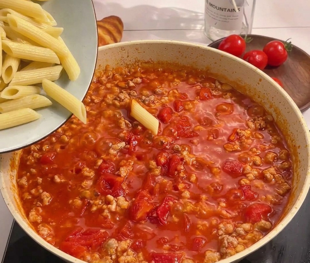 Gekochte Penne werden aus einer Schüssel in die Pfanne zur köchelnden Tomaten-Fleisch-Sauce gegeben.