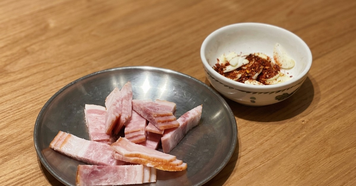 Roher Speck in Scheiben auf einem Metallteller neben einer kleinen Schüssel mit in Scheiben geschnittenem Knoblauch und Chiliflocken auf einem Holztisch.