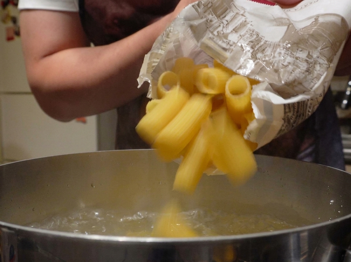Trockene Rigatoni-Nudeln fallen aus einer Papiertüte in einen Topf mit sprudelnd kochendem Wasser.