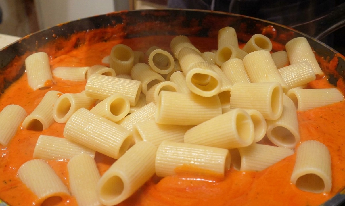 Gekochte Rigatoni-Nudeln werden in eine Pfanne gegeben, die mit einer glatten, cremigen, orange-roten Sauce gefüllt ist.