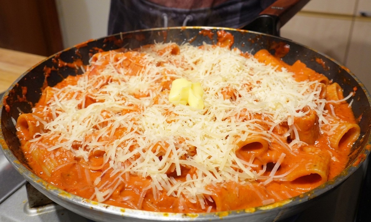 Ein kleiner Würfel gelber Butter ruht auf einem großzügigen Haufen von geriebenem Parmesan und Rigatoni-Nudeln in einer heißen Bratpfanne.