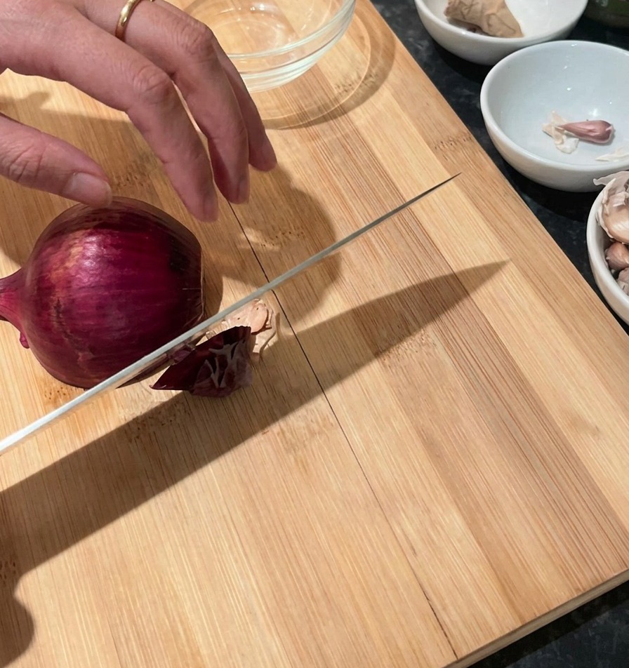 Eine Hand hält eine rote Zwiebel auf einem Holzschneidebrett, daneben liegt ein Kochmesser zum Schneiden bereit.