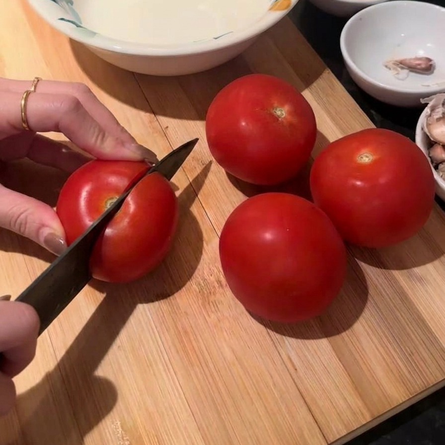 Hände schneiden eine frische rote Tomate mit einem Kochmesser auf einem Holzbrett, drei weitere Tomaten liegen daneben.