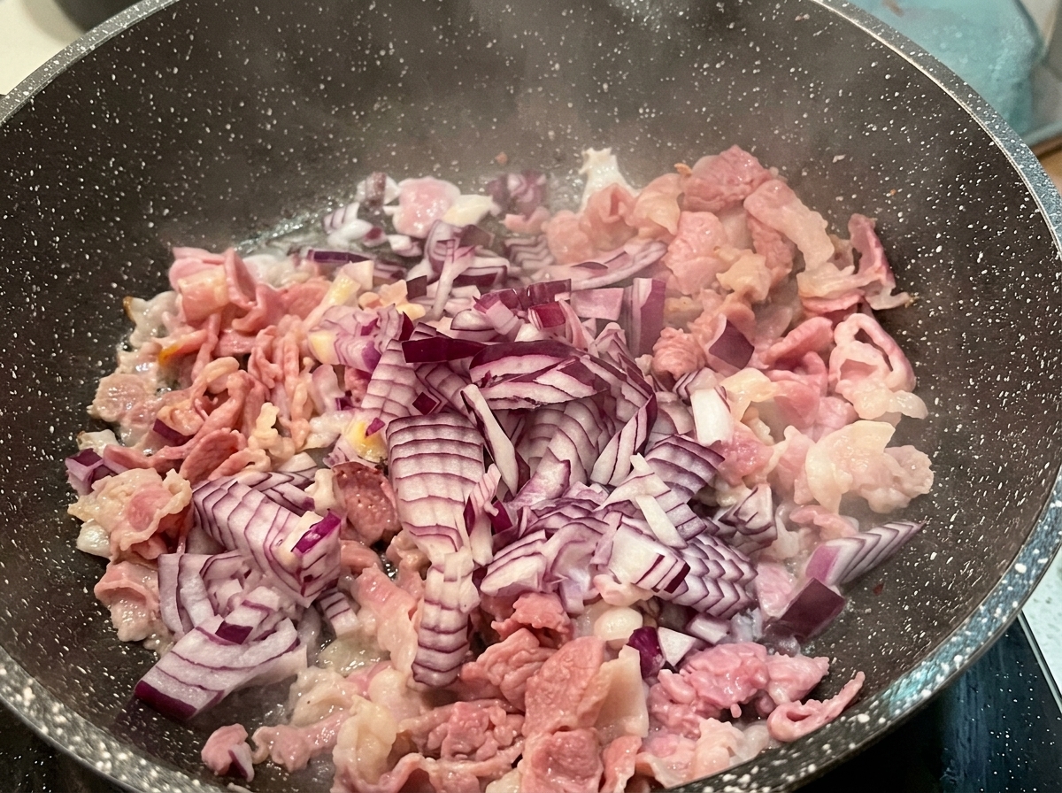 Gewürfelte rote Zwiebeln über teilweise gebratenem Speck in einer heißen, gesprenkelten Pfanne.