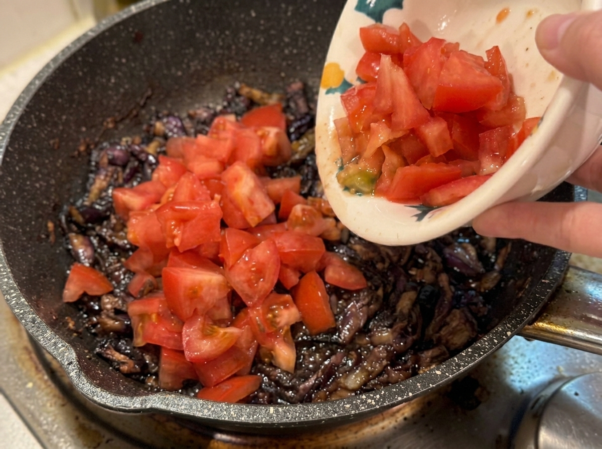 Eine Hand gießt gewürfelte Tomaten aus einer Keramikschüssel in eine heiße Pfanne mit Speck und Zwiebeln.