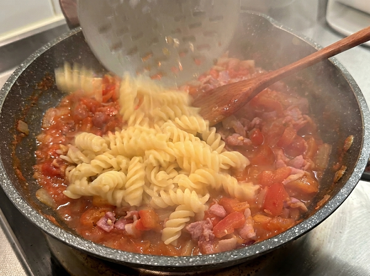 Gekochte Fusilli werden mit einem Schaumlöffel in die heiße rote Tomaten-Speck-Sauce gegeben.