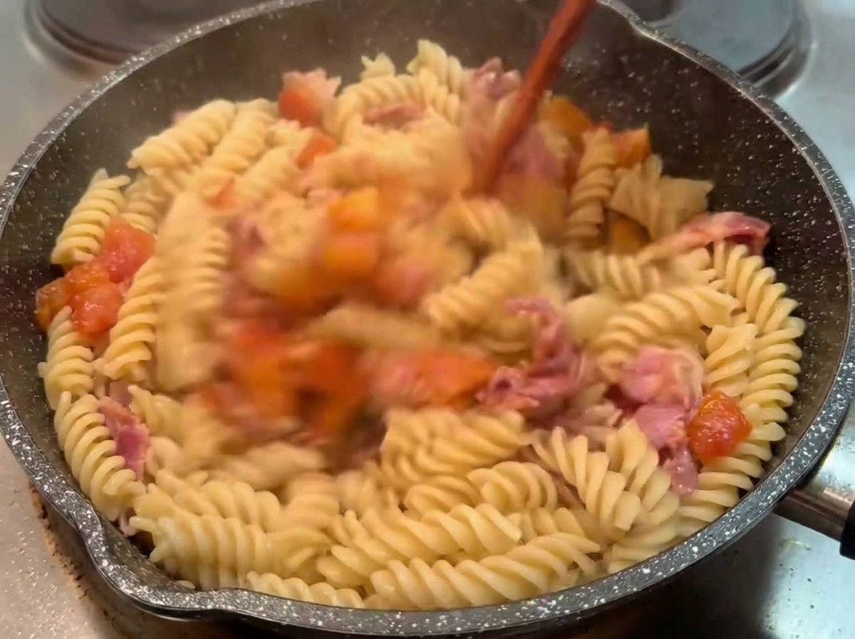 Gekochte Fusilli werden in einer dunklen Pfanne mit einem Holzlöffel unter die Tomaten-Speck-Sauce gemischt.