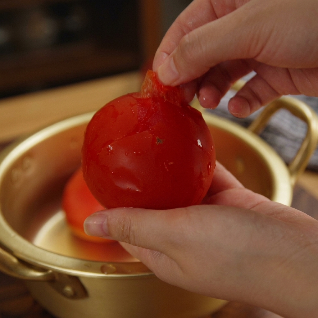 Hände ziehen die lose Haut von einer hellroten Tomate ab.