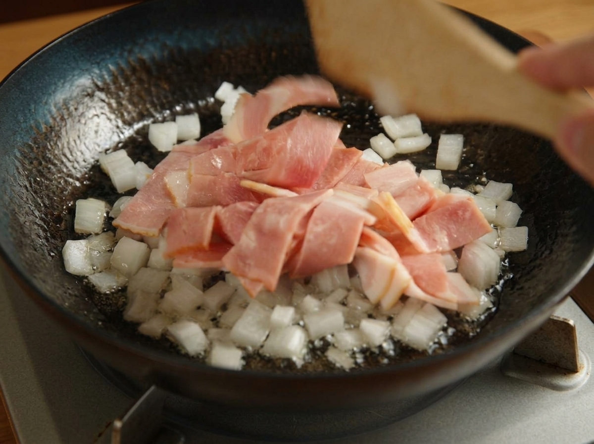 Speckstücke und Zwiebelwürfel werden in einer schwarzen Pfanne gebraten.