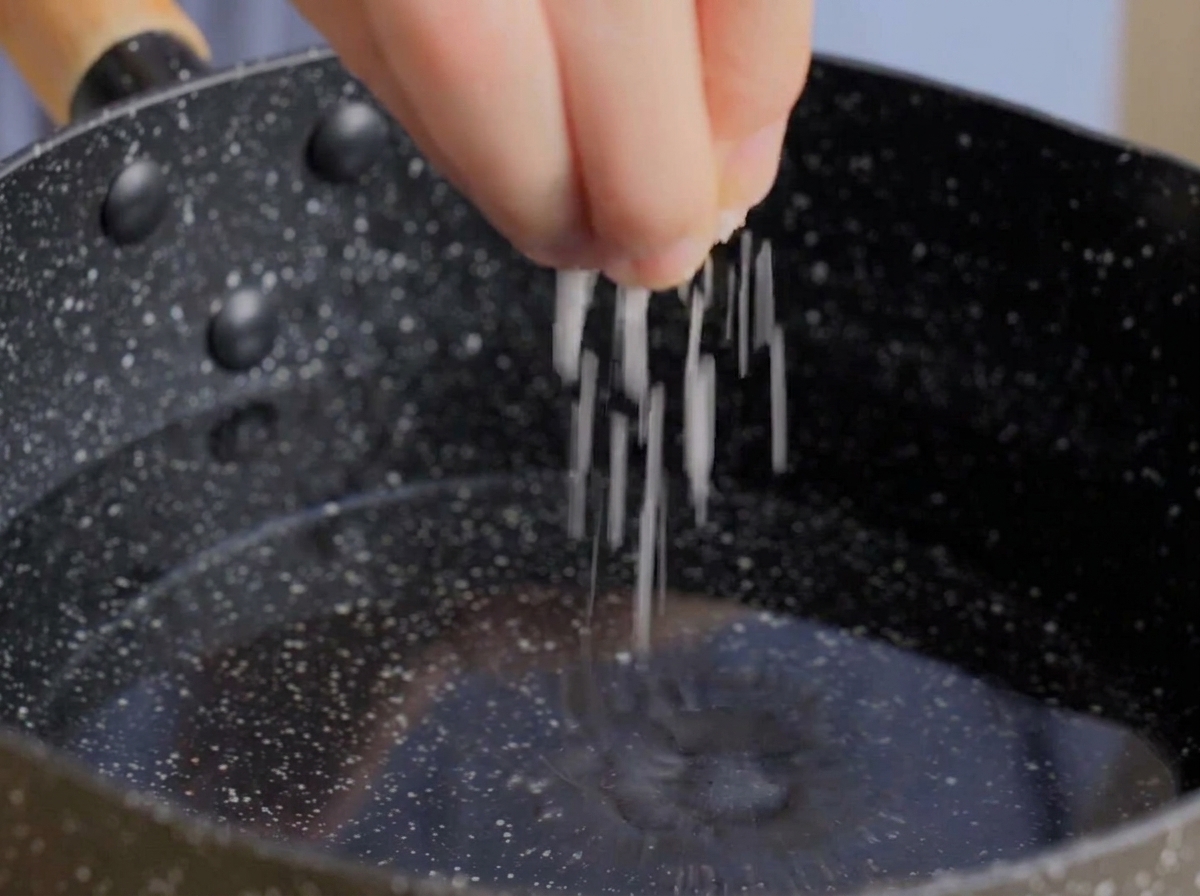 Eine Hand gibt eine Prise grobes Salz in einen dunklen Topf mit kochendem Wasser.