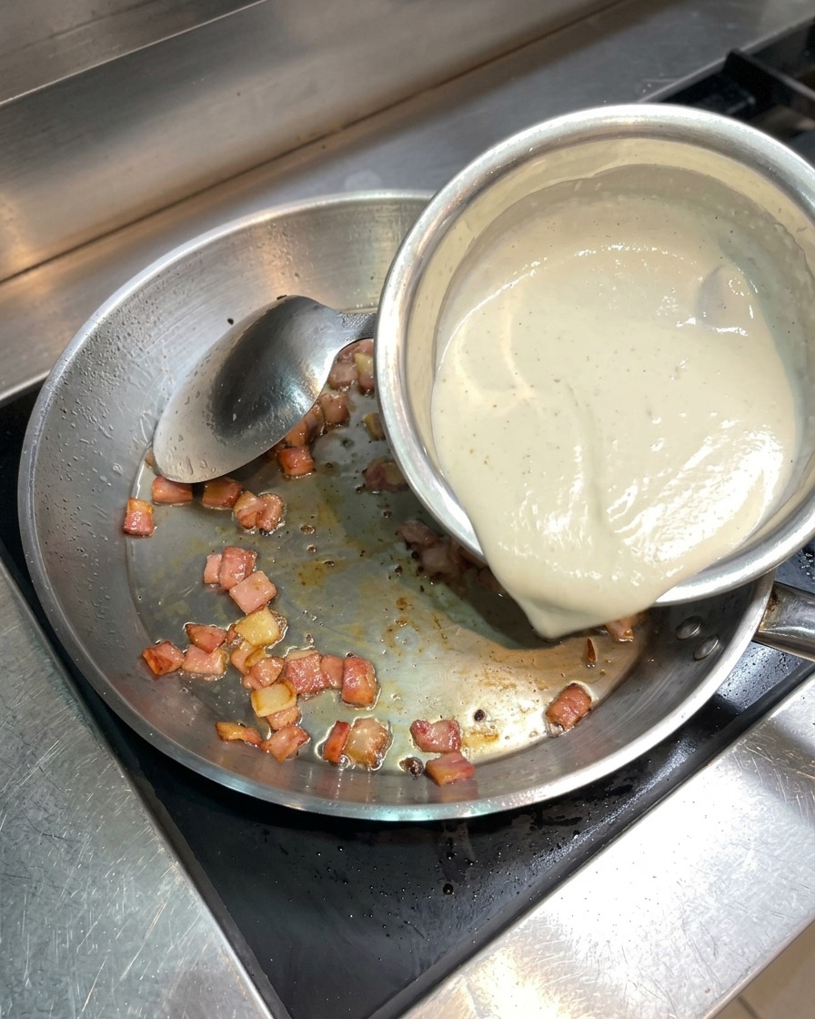 Flüssige Sahne wird aus einer kleinen Schüssel zu dem knusprigen Speck in die Pfanne gegossen.