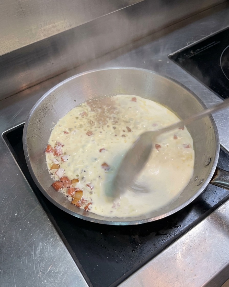 Eine Edelstahlpfanne mit köchelnder Sahne und Speck, die mit einem Metalllöffel umgerührt wird.