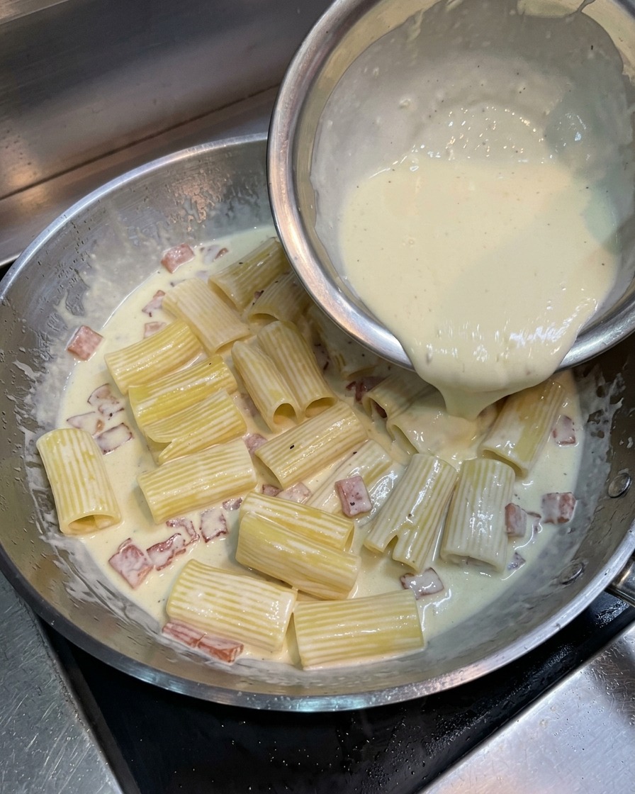 Eine hellgelbe Mischung aus Eigelb, Sahne und Käse wird über die Rigatoni in der Pfanne gegossen.