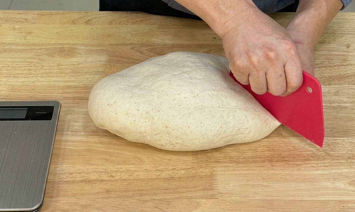 Ein Koch teilt den glatten Pizzateig mit einem Teigschaber auf einer Holzfläche.