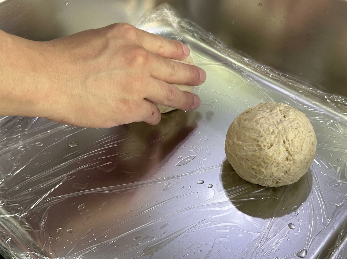 Geformte Teigballen auf einem Metallblech, mit Frischhaltefolie abgedeckt.
