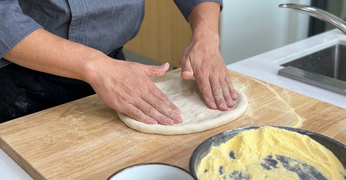Der Pizzateig wird auf einem Brett ausgebreitet, wobei ein dickerer Rand bleibt.