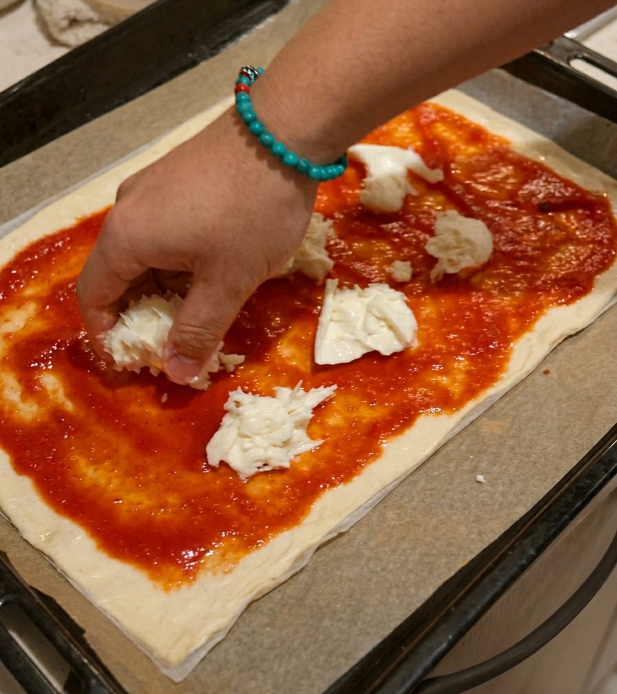 Eine Hand legt ungleichmäßig gezupfte Stücke weißen Mozzarellas auf die mit Tomatensauce bestrichene Pizza.