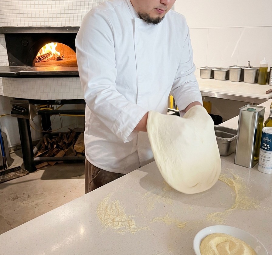 Ein Koch drückt und zieht einen runden Pizzateig auf einer bemehlten Arbeitsfläche.