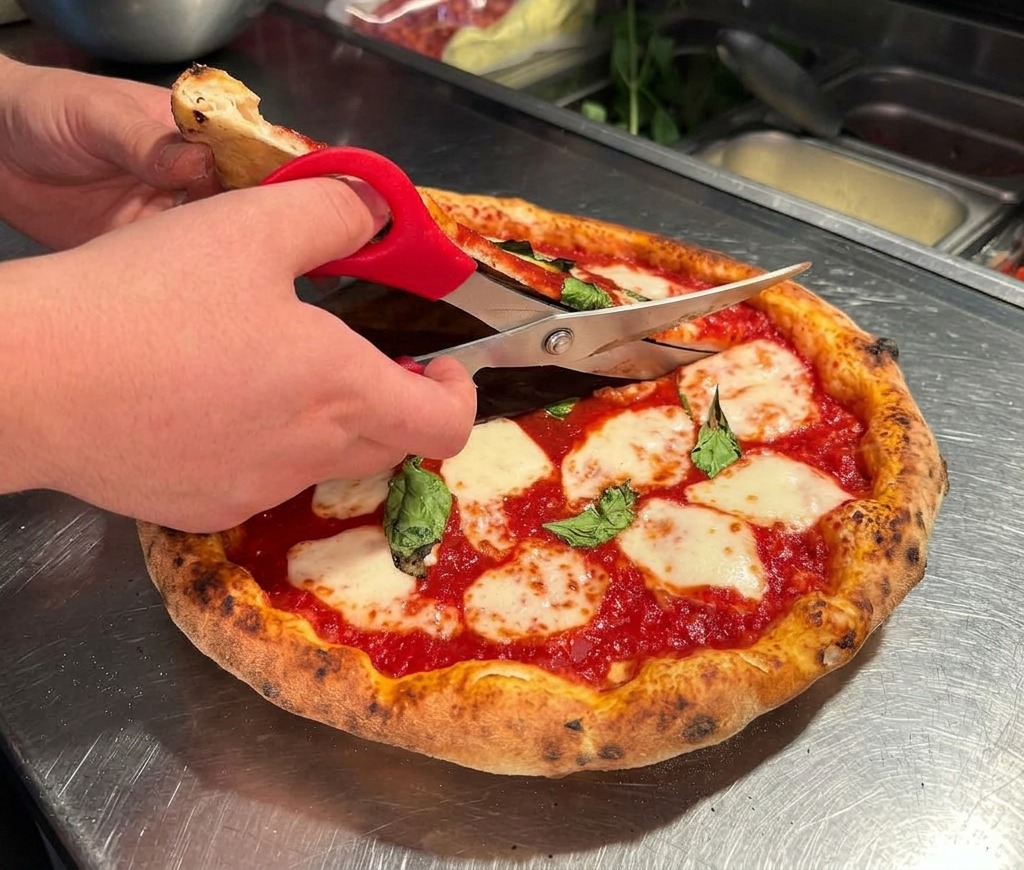 Hände schneiden die heiße Pizza mit einer Küchenschere auf einer Edelstahloberfläche.