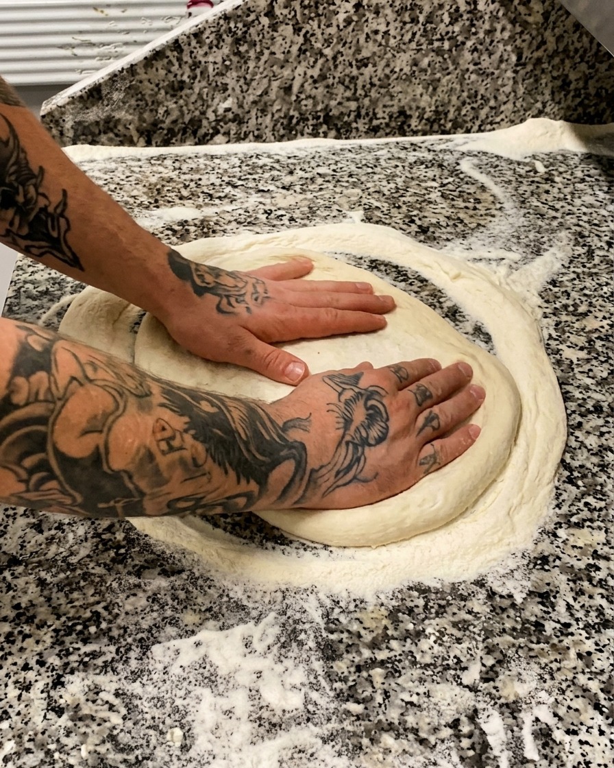 Tätowierte Hände drücken und dehnen einen runden Pizzateig auf einer bemehlten Marmorplatte.