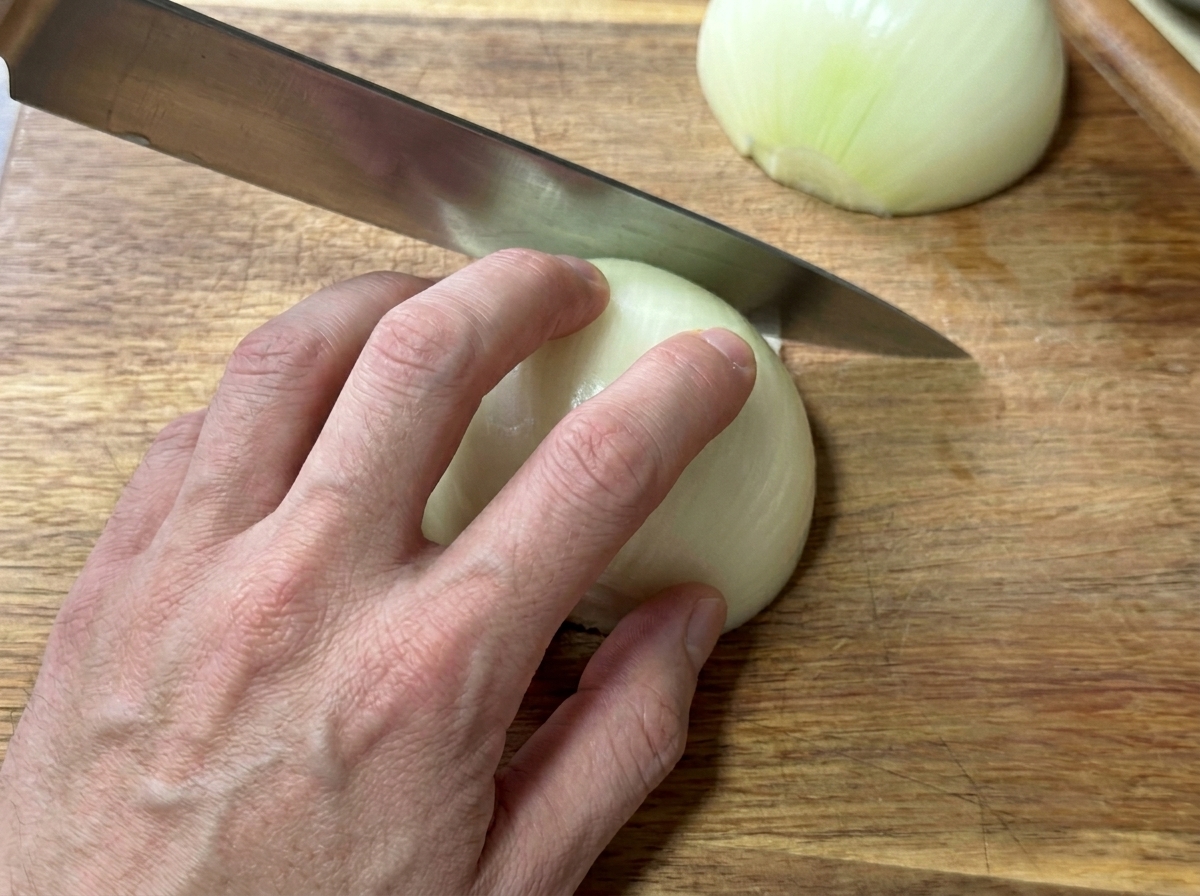 Eine Hand schneidet eine geschälte weiße Zwiebel auf einem Holzbrett in feine Streifen.