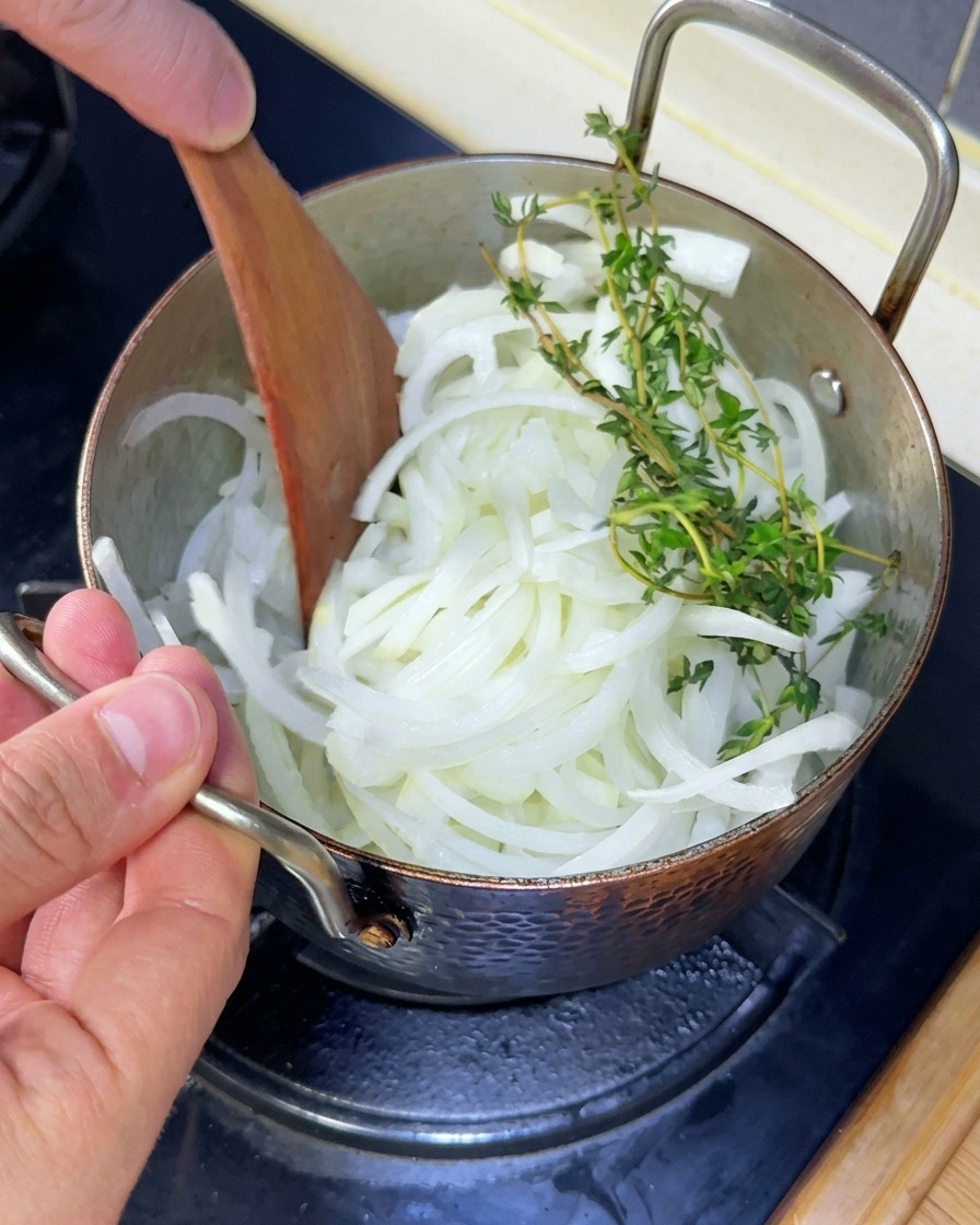 Weiße Zwiebelstreifen und frische Thymianzweige werden mit einem Holzspatel in einem Topf umgerührt.