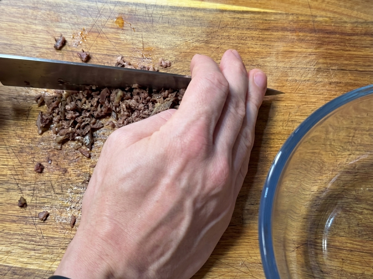 Eine Hand hackt Sardellenfilets mit einem großen Messer fein auf einem Holzbrett.