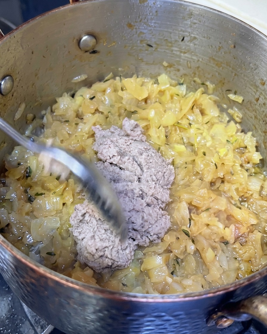 Ein Löffel der grauen Fischpaste wird zu den karamellisierten Zwiebeln in den Topf gegeben.