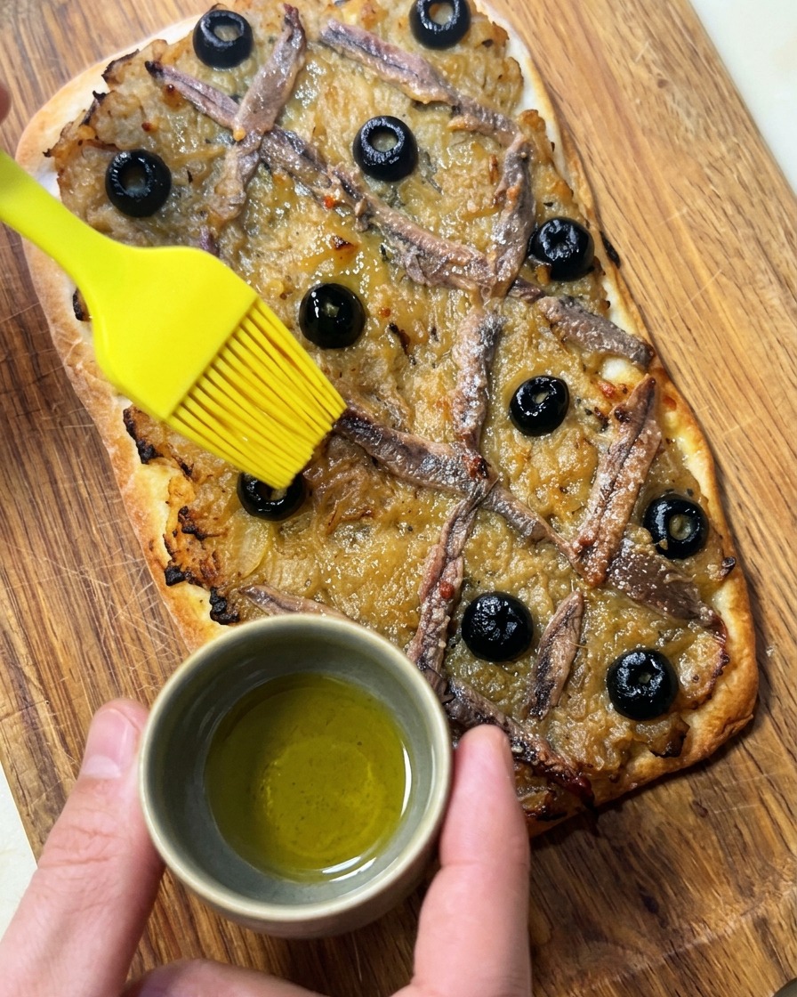 Ein Silikonpinsel bestreicht die fertig gebackene Pissaladière mit Olivenöl.