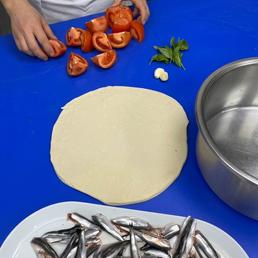 Ein Koch nimmt geviertelte Tomaten von der Arbeitsfläche, daneben liegen Knoblauch und frischer Basilikum.