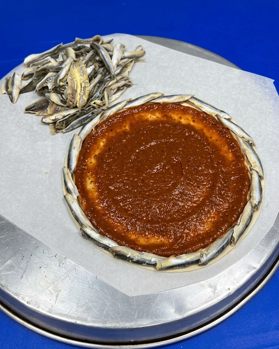 Ein runder Pizzaboden mit Sardellenrand, dessen Mitte gleichmäßig mit roter Tomatensauce bestrichen ist.