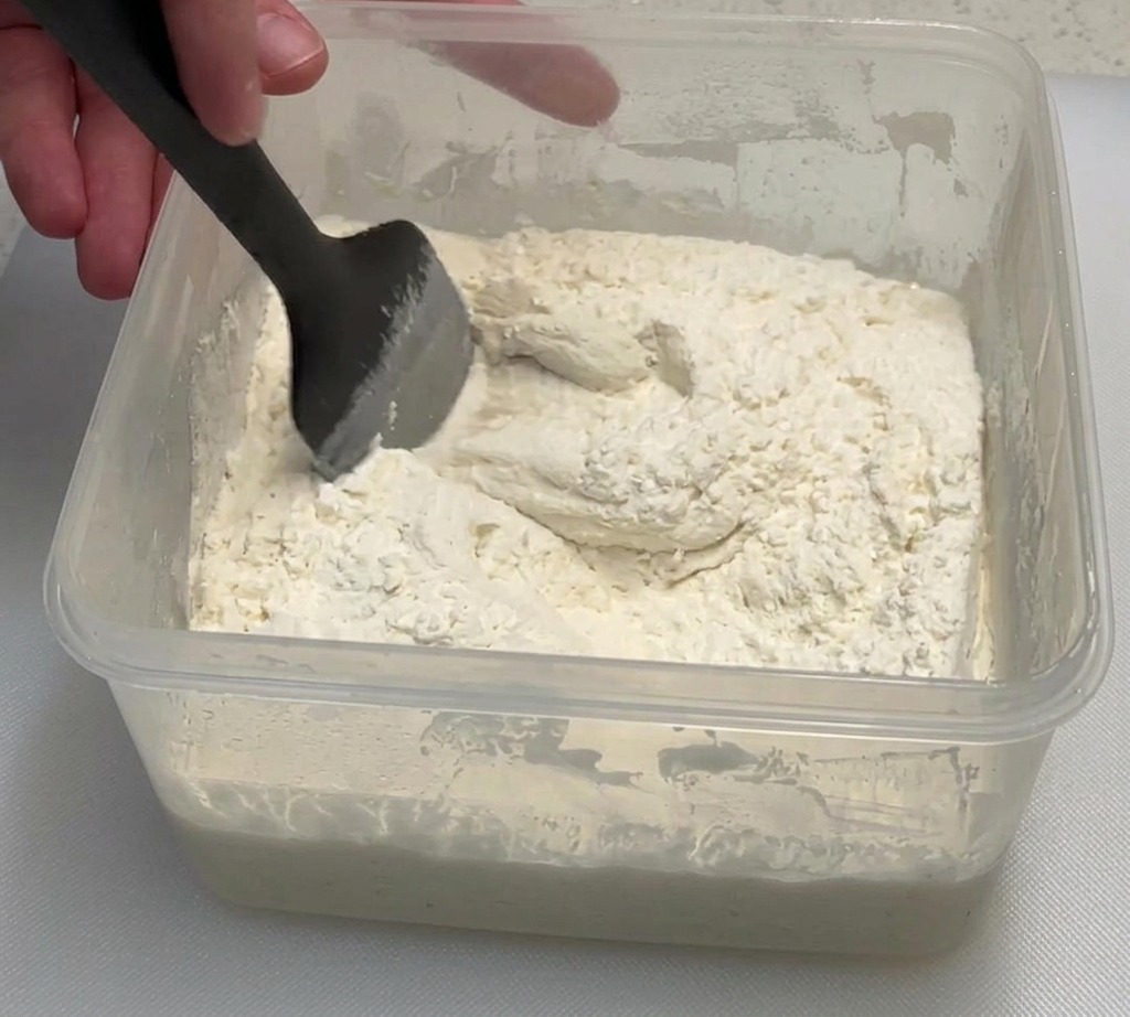 Eine Hand mischt mit einem schwarzen Spatel eine klebrige Mehl- und Poolish-Mischung in einem Klarsichtbehälter.