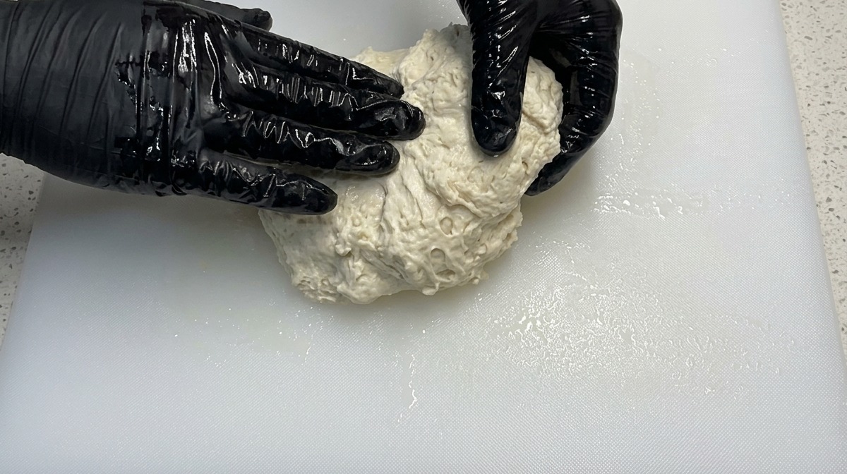 Hände in schwarzen Handschuhen kneten einen feuchten, klebrigen Pizzateig auf einem weißen Schneidebrett.