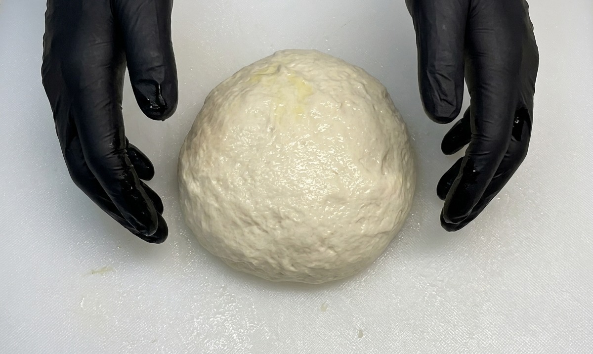 Hände in schwarzen Handschuhen formen eine glatte, runde Pizzateigkugel auf einem weißen Brett.