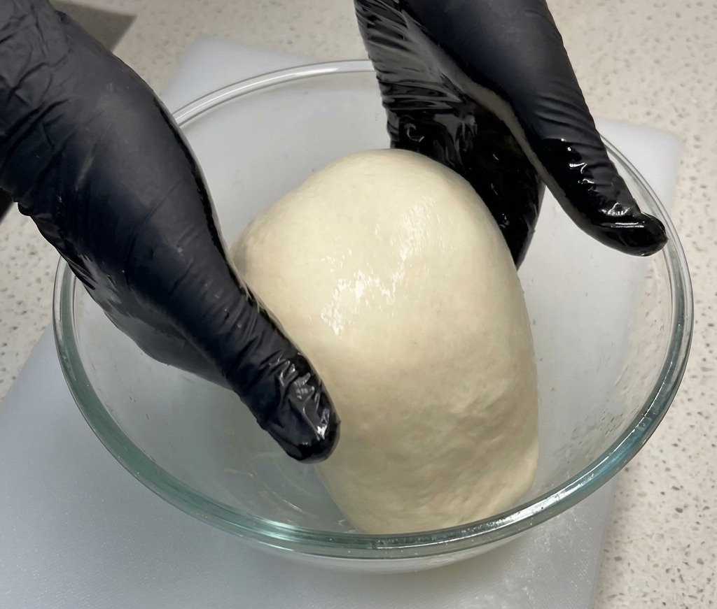 Handschuhe tragende Hände heben die Mitte eines weichen Pizzateigs in einer Glasschüssel vorsichtig nach oben.