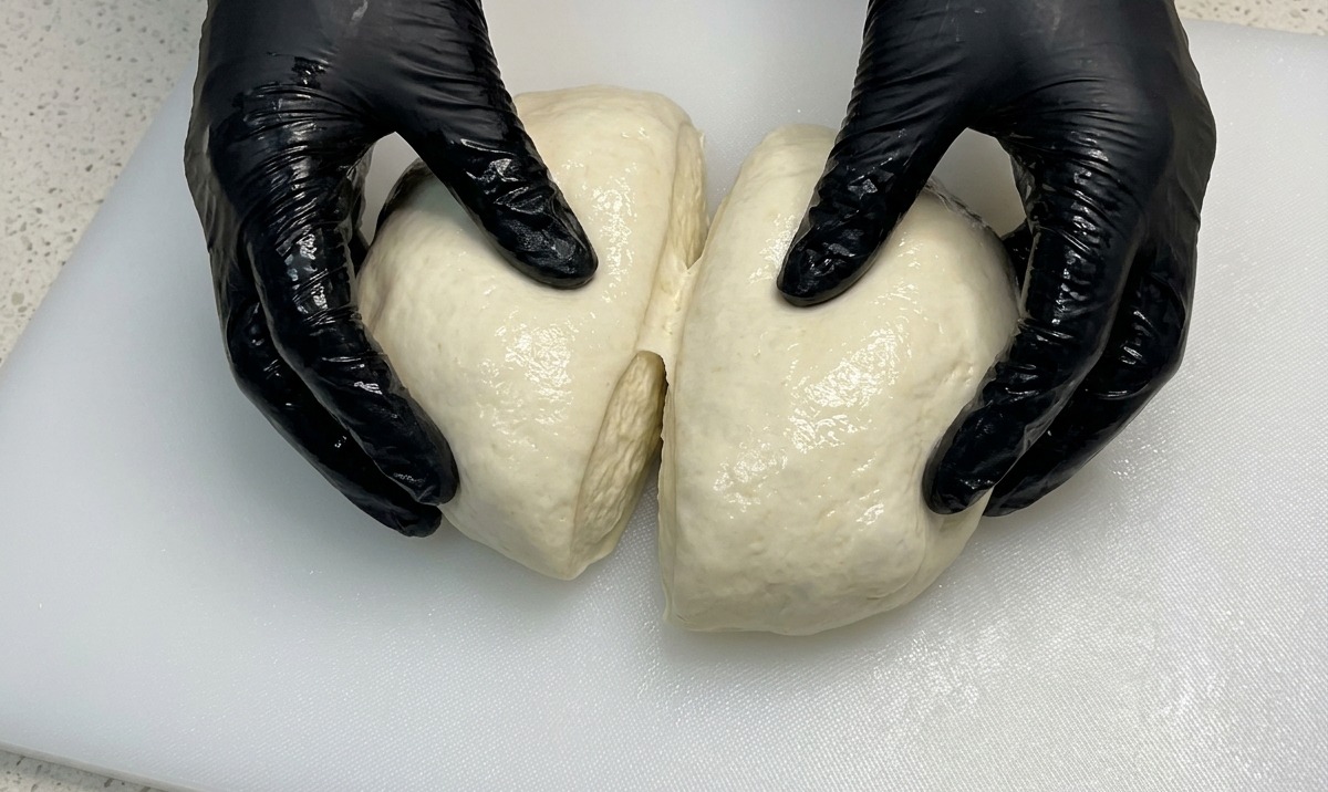 Hände in schwarzen Handschuhen teilen eine große, luftige Teigkugel in zwei gleiche Hälften.