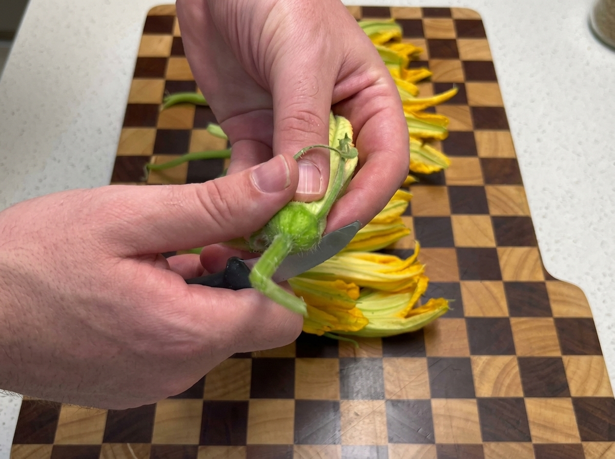 Hände schneiden mit einem kleinen Messer den Stiel von einer frischen gelben Kürbisblüte auf einem Holzbrett ab.