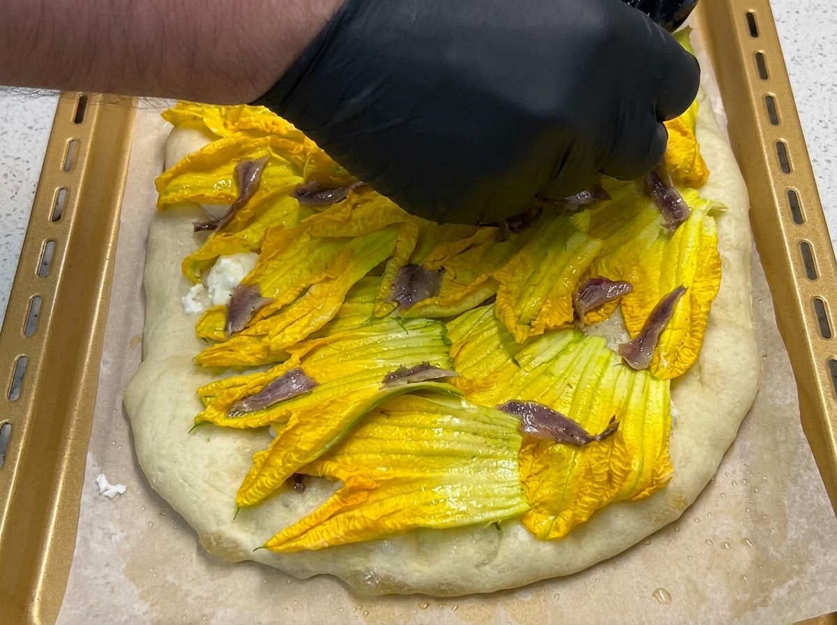 Eine Hand verteilt kleine dunkle Sardellenfilets oben auf den gelben Kürbisblüten auf der Pizza.