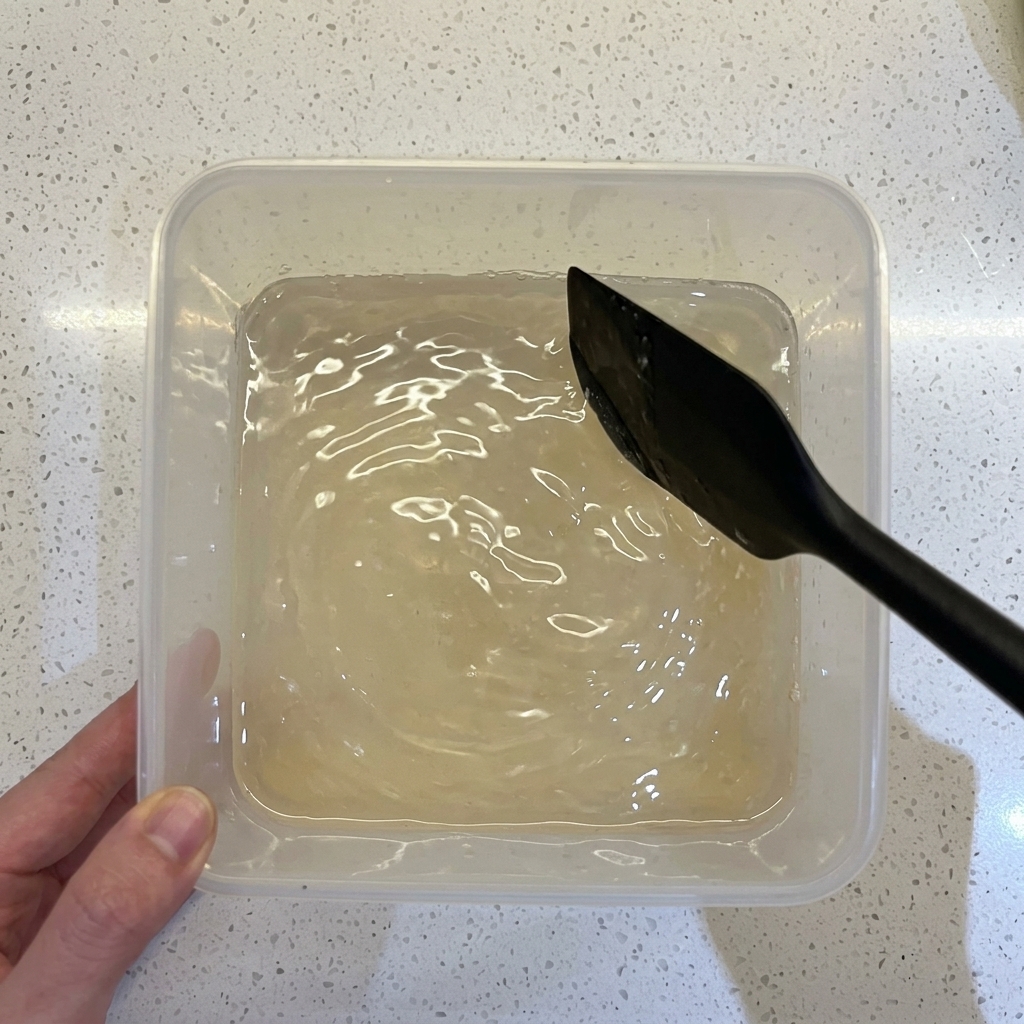 Ein schwarzer Spatel rührt eine wässrige Mehl-Hefe-Mischung in einem transparenten Behälter um.