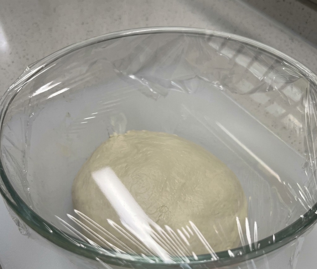 Eine glatte Pizzateigkugel ruht in einer großen Glasschüssel, die fest mit Frischhaltefolie verschlossen ist.