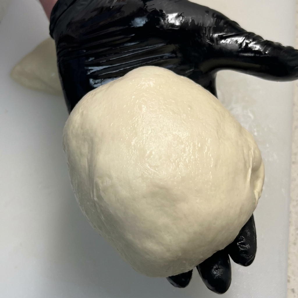 Eine glänzende, perfekt geformte Teigkugel liegt in der Handfläche eines schwarzen Handschuhs.