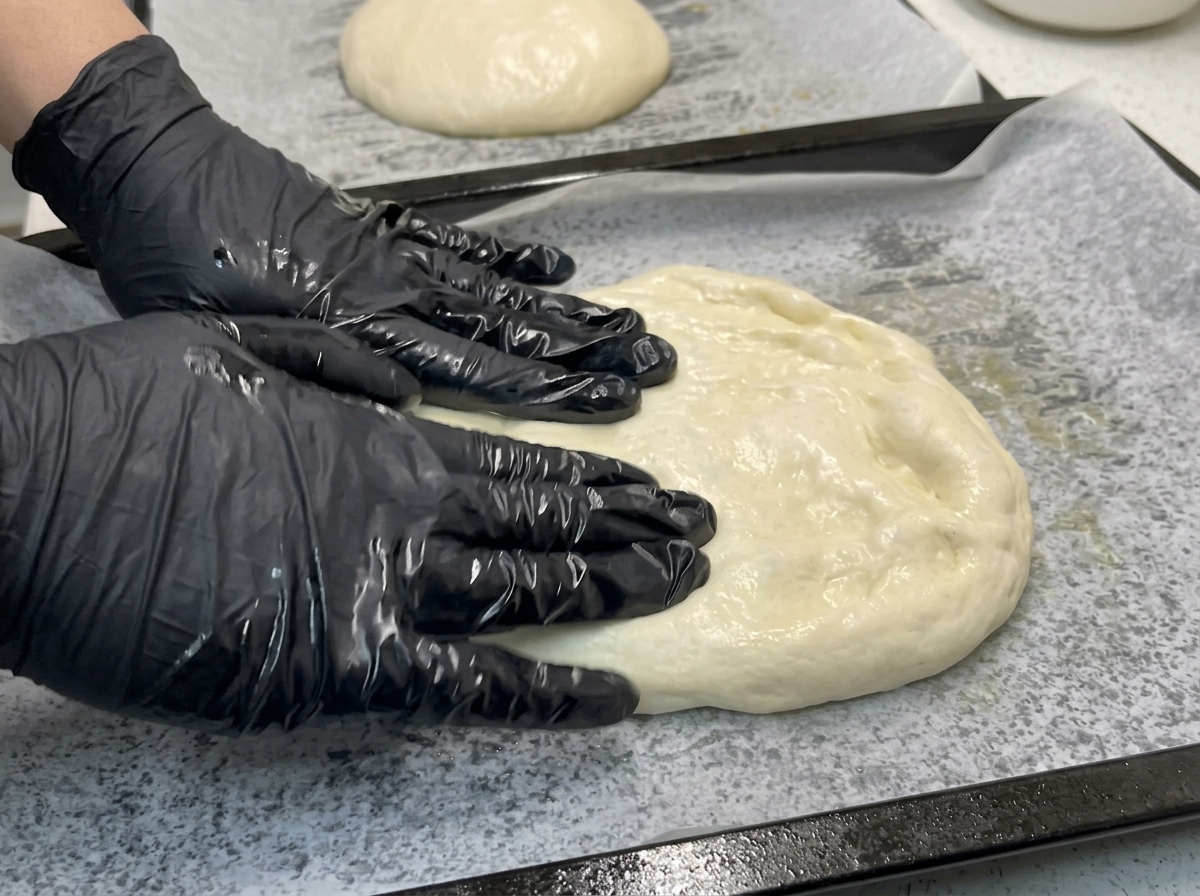 Hände in Handschuhen drücken und dehnen einen weichen Teigfladen auf einem Backblech mit Backpapier.
