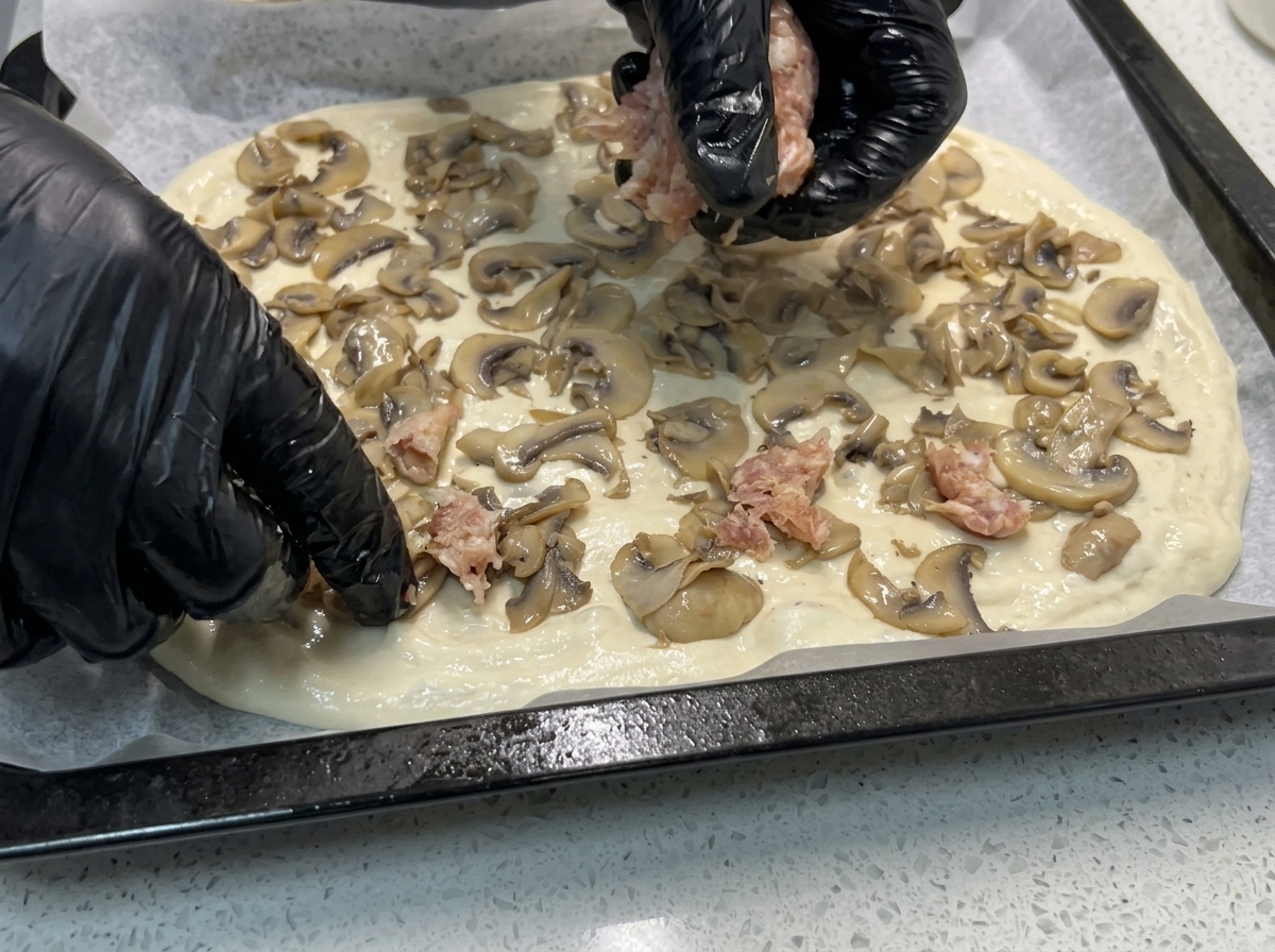 Hände verteilen Stücke von rohem Salsiccia-Brät auf den mit Pilzen belegten Pizzateig.