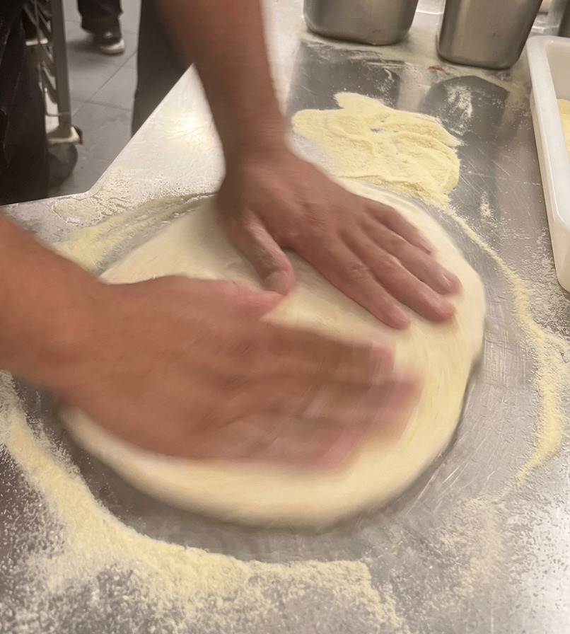 Hände dehnen den Pizzateig auf einer Edelstahlarbeitsplatte zu einem Kreis.