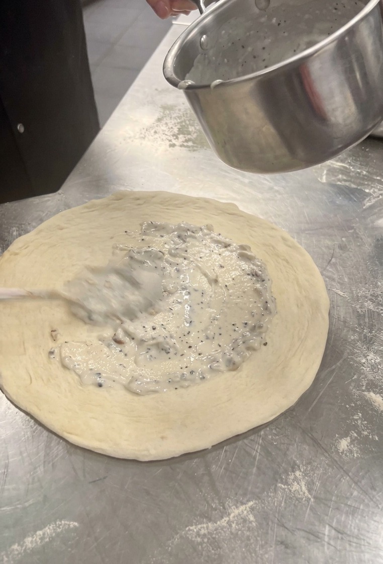 Cremige weiße Pilzsauce wird mit einem Löffel auf dem Pizzateig verteilt.