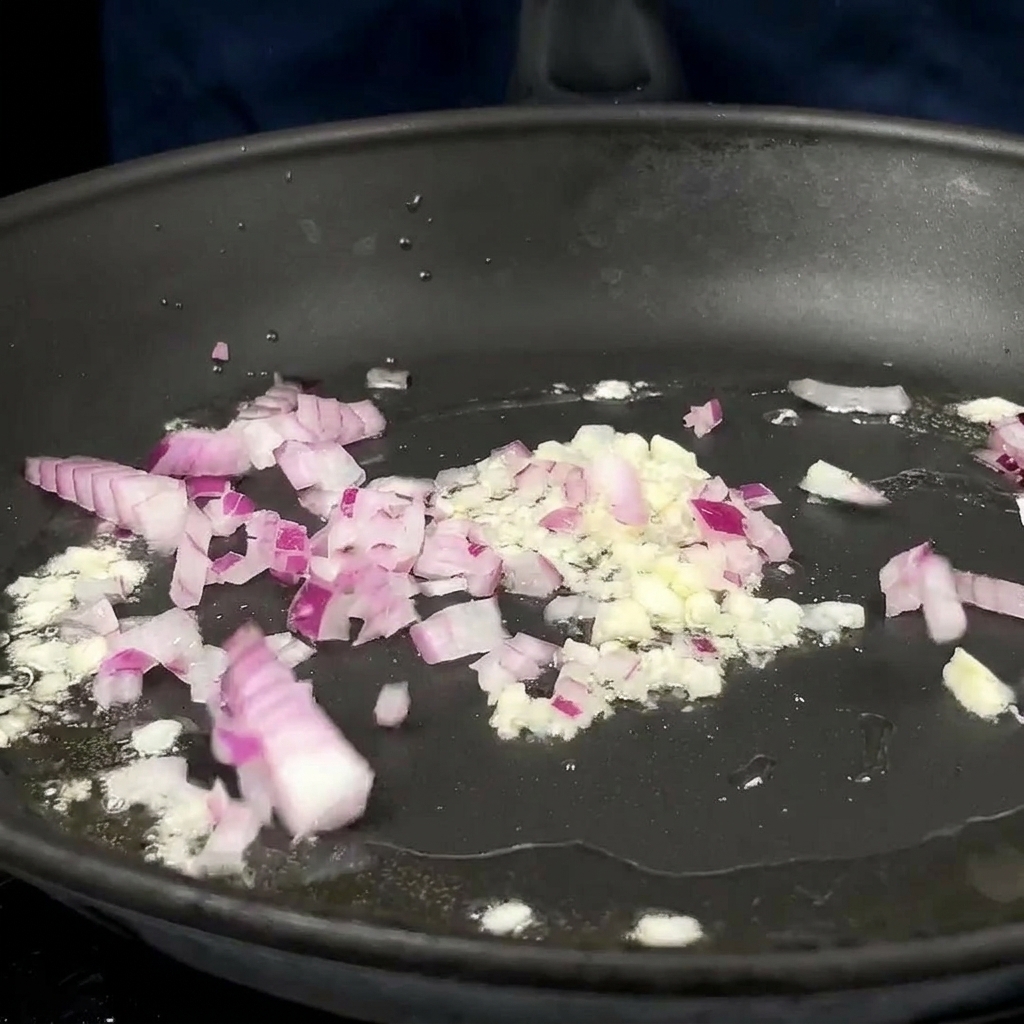 Gewürfelte rote Zwiebeln und gehackter Knoblauch brutzeln in heißem Öl in einer dunklen Pfanne.