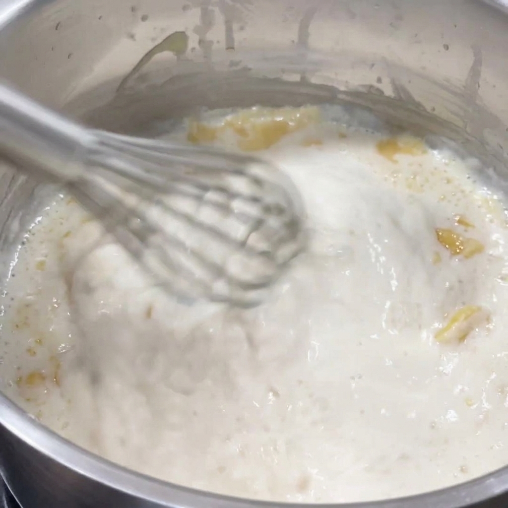 Ein Schneebesen rührt eine dicke, blubbernde weiße Sauce in einem Edelstahltopf auf.