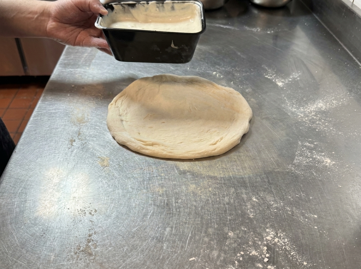 Trüffelsauce wird auf einen runden, rohen Pizzateig auf einer Edelstahloberfläche gegeben.