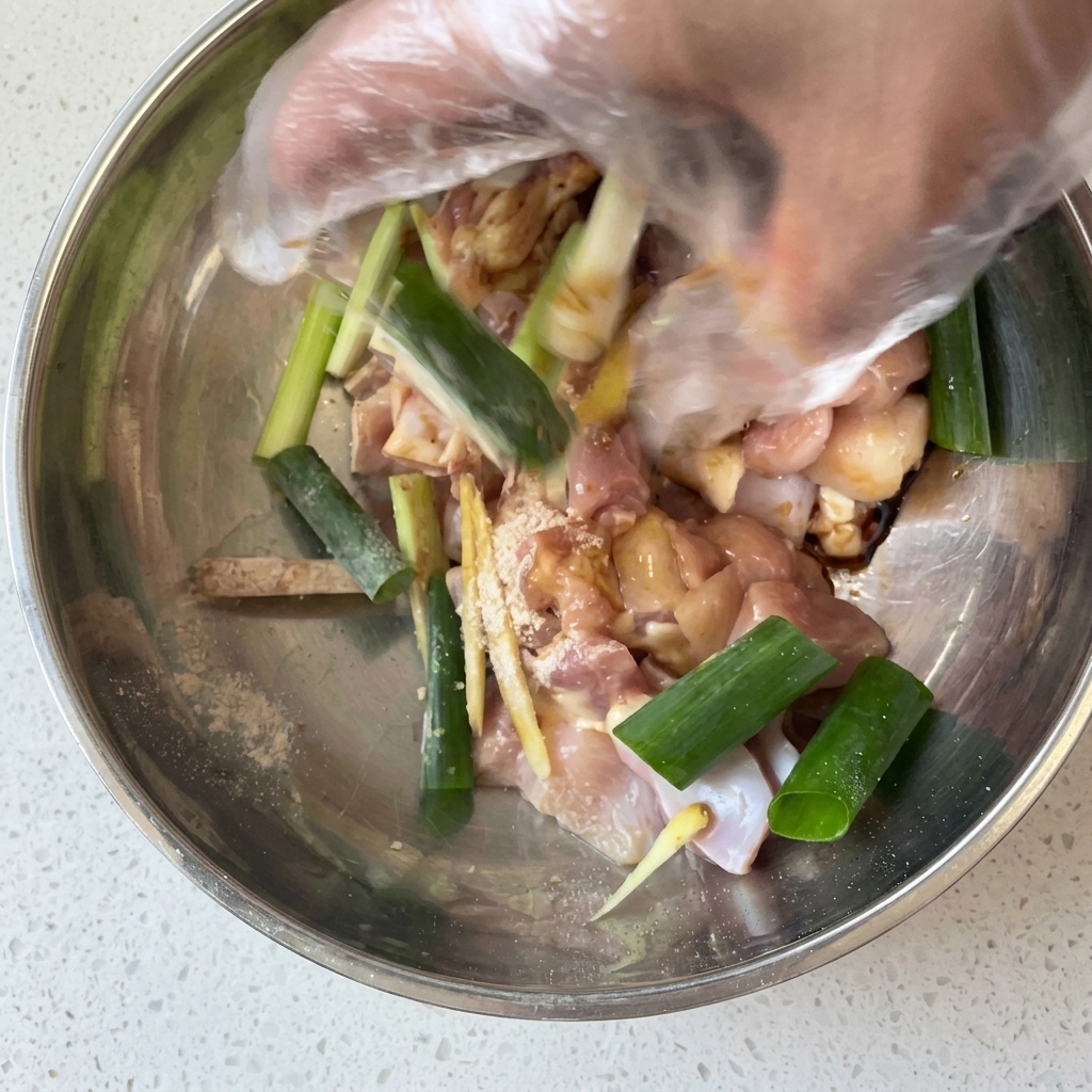 Eine Hand mit Plastikhandschuh mischt rohe Hähnchenstücke mit Frühlingszwiebeln und Marinade in einer Schüssel.