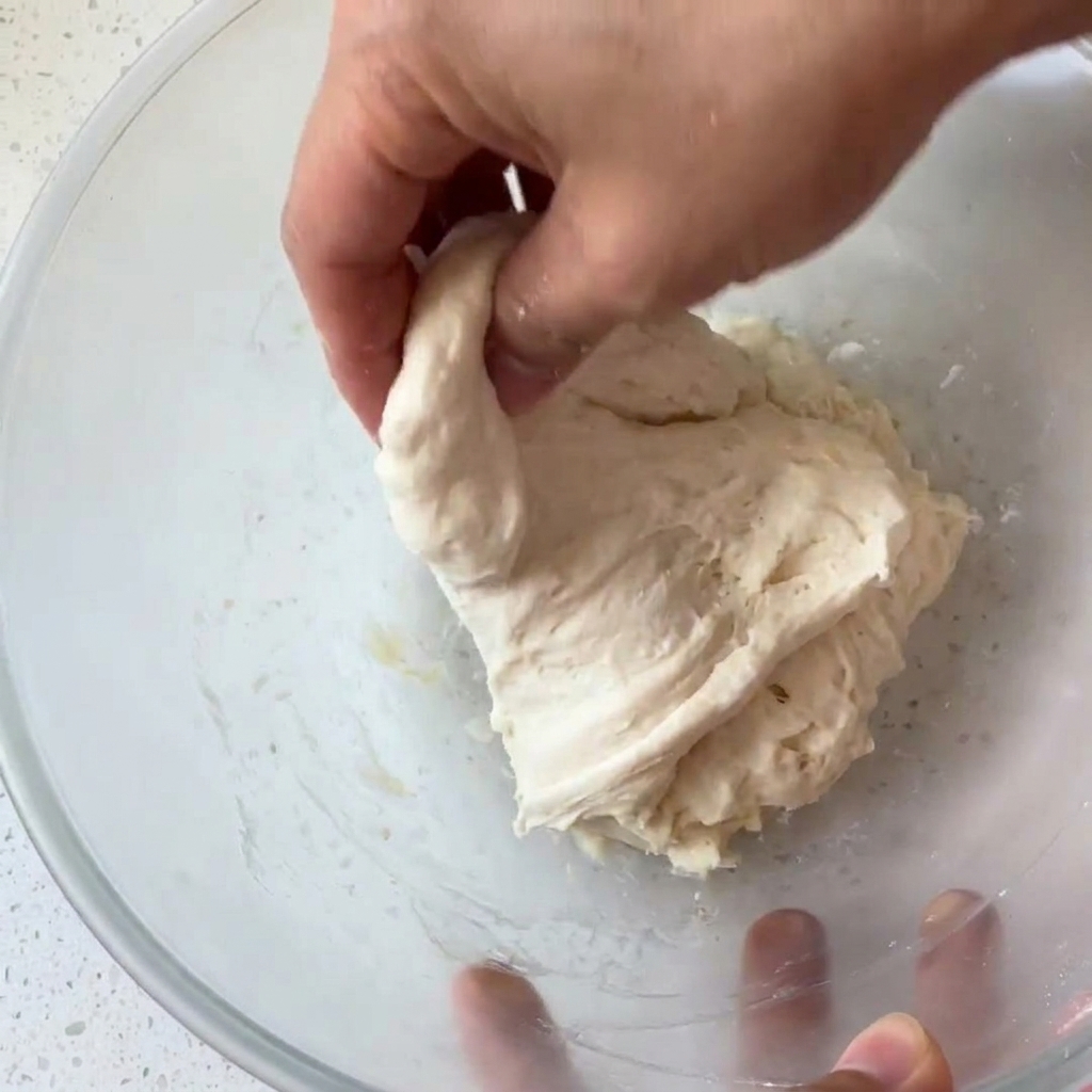 Eine Hand greift und faltet einen feuchten, klebrigen Teigballen in einer Glasschüssel.